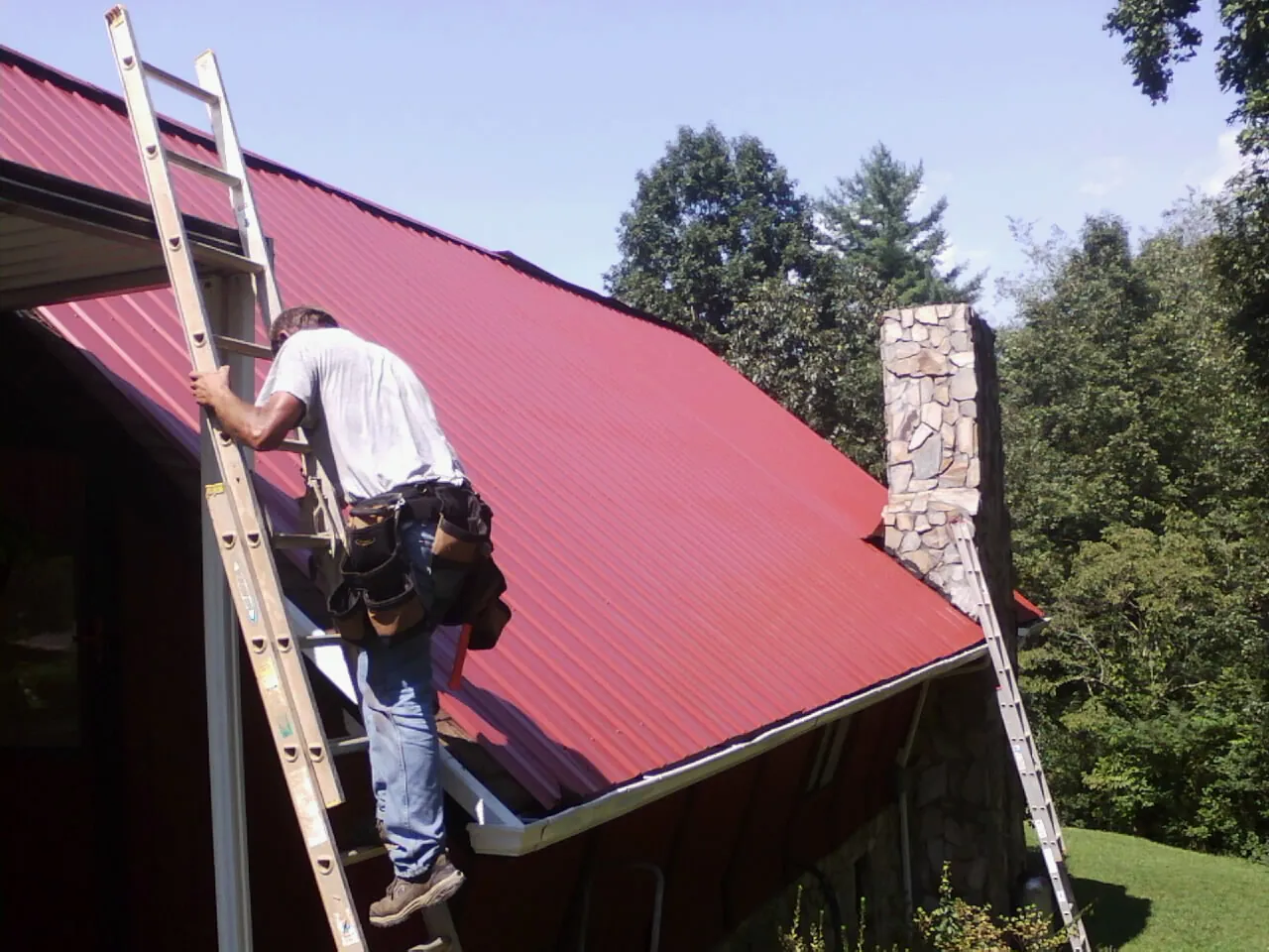 Roofer on a ladder beside a red metal roof and stone chimney — Soffit & Fascia Repair by Roofing Brothers in White Salmon